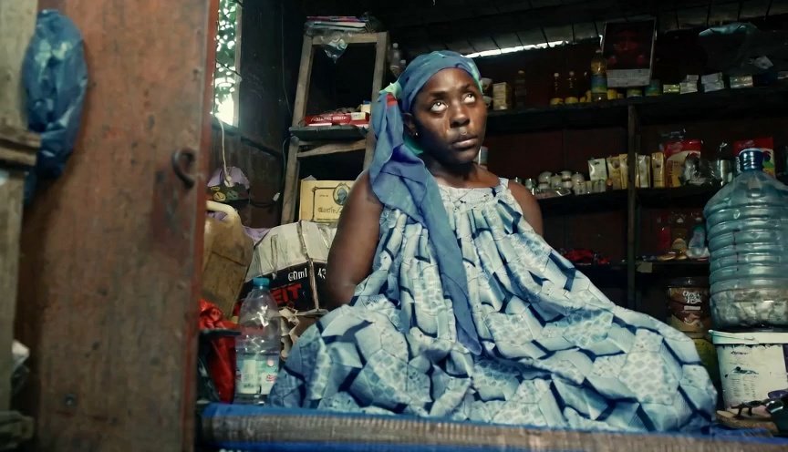 A woman in a blue dress sits inside a small shop, looking upward